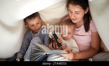 Deux garçons en pyjama écoutant l'histoire de conte de fées de mère tout en se cachant sous couverture la nuit. Famille ayant du temps ensemble, parentalité, enfant heureux Banque D'Images