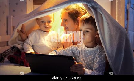 Mère souriante avec deux garçons assis dans une tente de couverture dans le lit et utilisant un ordinateur tablette. Famille ayant du temps ensemble, enfants avec des gadgets, parentalité, Banque D'Images