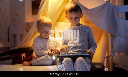 Deux garçons en pyjama jouant sur le lit la nuit et utilisant une tablette. Banque D'Images