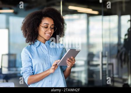 Jeune belle femme réussie au lieu de travail à l'intérieur du bureau avec tablette dans les mains, programmeuse femme testant la nouvelle application, souriant heureux. Banque D'Images