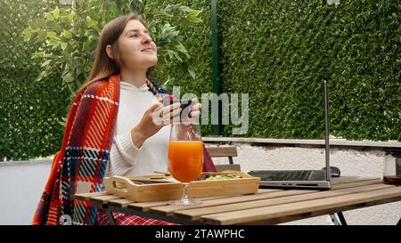 Jeune femme couverte de plaid travaillant sur ordinateur portable à la terrasse extérieure et buvant du café. Concept de freelance, travail à domicile, travail à distance, eatin Banque D'Images