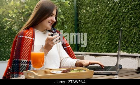 Jeune femme souriante couverte de plaid boire du café sur la terrasse et boire du thé ou du café tout en travaillant sur ordinateur portable. Concept de freelance, travail Banque D'Images