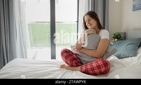 Jeune femme rêveuse en pyjama confortable assise sur le lit, serrant un coussin et souriant à la pensée de sa bien-aimée Banque D'Images