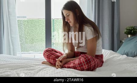 Bouleversée et solitaire jeune femme en pyjama assise sur le lit à côté de la grande fenêtre. Banque D'Images