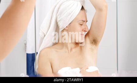 Jeune femme sentant ses aisselles malodorantes avec des cheveux qui poussent à la salle de bain. Concept d'hygiène, de beauté naturelle, de féminité et de croissance des poils du corps Banque D'Images