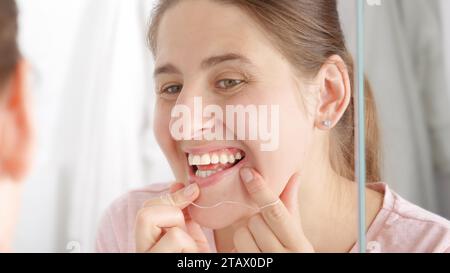 Portrait en gros plan d'une jeune femme vérifiant ses dents au miroir et utilisant du fil dentaire. Concept de santé dentaire, autocontrôle buccal et hygiène bucco-dentaire Banque D'Images