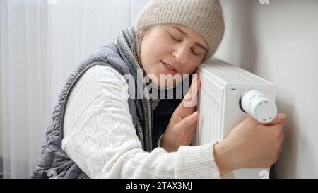 Jeune femme en écharpe et chapeau à la maison embrassant radiateur de chauffage. Concept de crise énergétique, factures élevées, système de chauffage cassé, économie et économie d'argent Banque D'Images