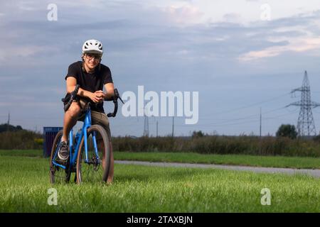 Heureux cycliste masculin portant casque et lunettes se tient avec son vélo de gravier sur l'herbe verte dans le parc. Banque D'Images