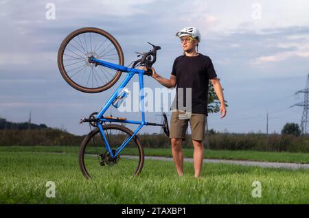 Heureux cycliste masculin portant casque et lunettes tient son vélo de gravier sur l'herbe verte dans le parc. Concept d'activités sportives. Banque D'Images