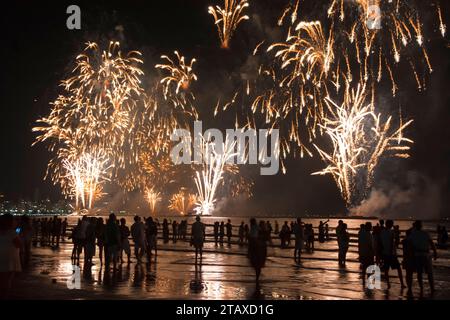 Plusieurs personnes à une célébration du nouvel an dans la ville avec des feux d'artifice sur la plage. Silhouettes et reflets dans l'eau. Banque D'Images