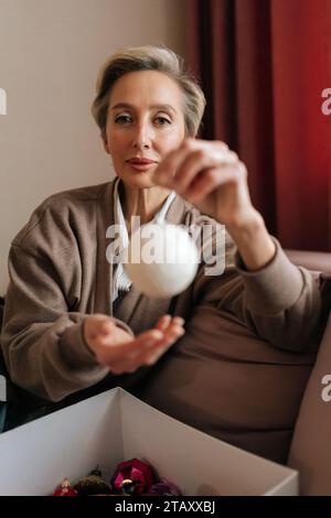 Portrait vertical d'une femme d'âge moyen tenant une boule de Noël, regardant la caméra avec une expression heureuse. La femelle élégante décore l'arbre de Noël pour New Banque D'Images