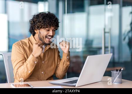 Homme d'affaires gagnant au lieu de travail à l'intérieur du bureau, l'homme a reçu un message en ligne sur les résultats réussis réalisation financière, travailler avec un ordinateur portable, tenant les mains vers le haut geste de triomphe. Banque D'Images