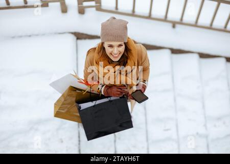 Vue supérieure de la femme heureuse et élégante d'âge moyen dans le chapeau brun et écharpe en manteau de chameau avec des gants et des sacs à provisions en utilisant des applications smartphone à l'extérieur Banque D'Images
