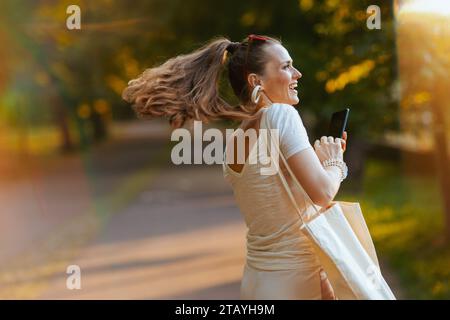 Heure d'été. souriant femme tendance en chemise blanche avec smartphone et sac fourre-tout marchant dans le parc de la ville. Banque D'Images