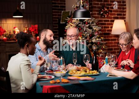 Famille heureuse sur les vacances de la veille de noël, rassemblement pour célébrer le dîner festif ensemble et boire de l'alcool, saison de décembre. Les jeunes et les seniors se sentent joyeux autour de la table avec de la nourriture et du vin. Banque D'Images