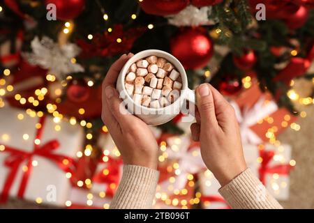 Femme tenant une tasse de délicieux cacao avec des guimauves près de l'arbre de Noël, vue de dessus Banque D'Images