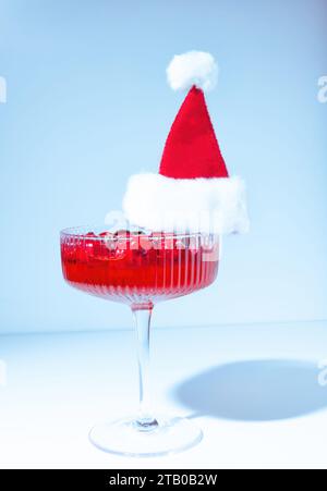 Un cocktail de canneberges rouges des fêtes dans un verre de champagne coupe W boucle d'oreille un chapeau de Père Noël rouge furry sur un fond bleu pastel Banque D'Images