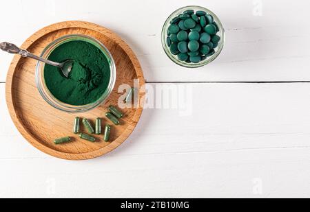 Poudre naturelle d'algues Spiruline verte organique dans un pot en verre, comprimés ronds, Capsules sur plateau rond en bois et fond blanc. Vue de dessus Banque D'Images