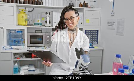Confiante et souriante, jeune scientifique hispanique, travaille magnifiquement avec le presse-papiers en laboratoire, immergé dans la recherche médicale Banque D'Images