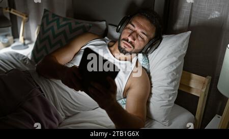 Jeune homme arabe utilisant le touchpad allongé sur le lit portant des écouteurs dans la chambre Banque D'Images