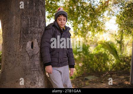 Un jeune garçon contemplatif se tient à côté d'un arbre, vêtu de vêtements d'hiver confortables, immergé dans l'ambiance sereine d'un parc ensoleillé. Banque D'Images