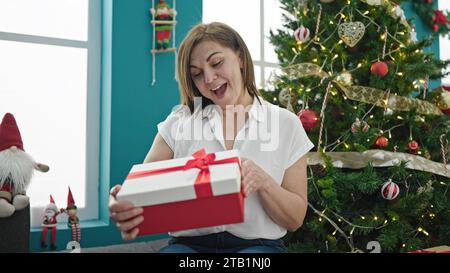 Femme hispanique d'âge moyen déballant cadeau avec visage surpris à la maison Banque D'Images