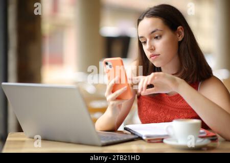 Femme sérieuse vérifiant téléphone et ordinateur portable dans une terrasse de bar Banque D'Images