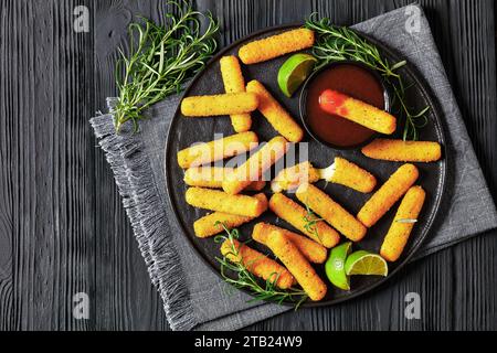 bâtonnets de mozzarella panés et frits servis avec sauce, tranches de citron vert et romarin frais sur assiette noire sur table en bois noir, cuisine américaine, Banque D'Images