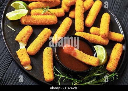 bâtonnets de mozzarella avec sauce, tranches de citron vert et romarin frais sur plateau noir sur table en bois noir, gros plan Banque D'Images