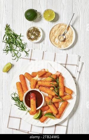 bâtonnets de mozzarella panés frits servis avec sauce, tranches de citron vert et romarin frais sur plateau blanc sur table en bois blanc avec ingrédients, ver Banque D'Images