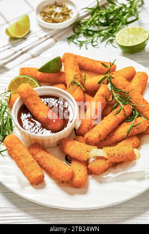 bâtonnets de mozzarella panés frits servis avec sauce, tranches de citron vert et romarin frais sur assiette blanche sur table en bois blanc avec ingrédients, verti Banque D'Images