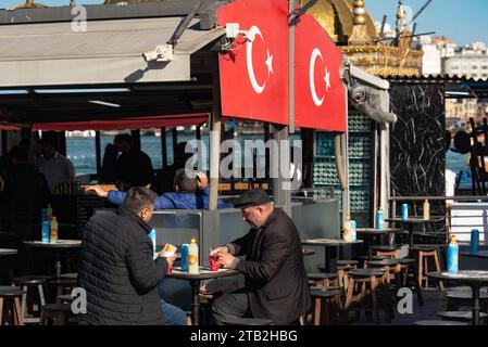 Istanbul, Turquie. 21 novembre 2023 deux hommes turcs mangent Balik Ekmek, un sandwich au poisson, un aliment de rue populaire vendu à côté de la Corne d'Or et de Bos Banque D'Images