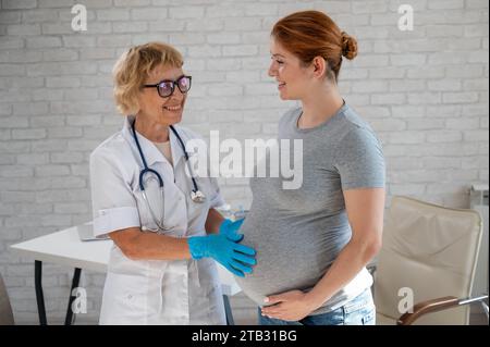 Femme enceinte consultant un médecin. Gynécologue caucasienne âgée tient les mains sur le ventre d'une patiente enceinte. Banque D'Images