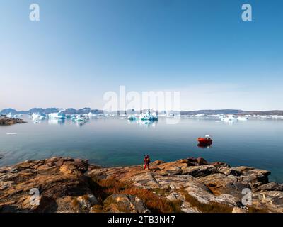 Un homme se tient sur un fjord groenlandais avec un bateau et des icebergs. Banque D'Images