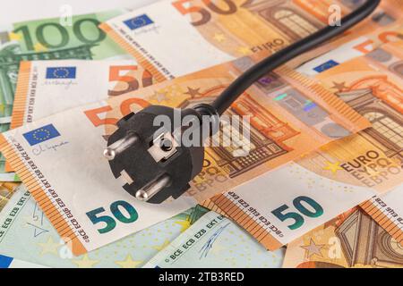 Une fiche électrique noire sur une table recouverte d’une grande quantité de billets en euros sur fond clair en gros plan Banque D'Images