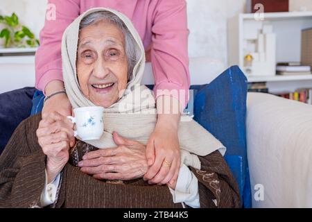 Les mains féminines embrassent sa grand-mère tout en buvant du café dans le salon sur le canapé Banque D'Images