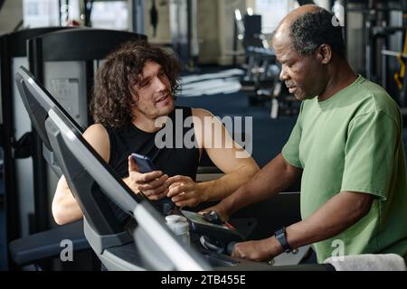 Jeune sportif caucasien parlant à un joueur de gym noir âgé, debout sur un tapis roulant au centre de fitness Banque D'Images