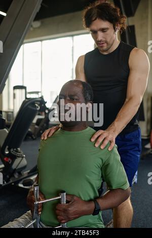 Homme noir senior tirant du poids à la salle de gym avec les conseils d'un jeune instructeur caucasien debout derrière Banque D'Images