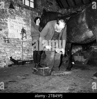 Années 1970, historique, à l'intérieur d'une vieille grange en pierre, un maréchal ferrant un cheval, avec une fille d'écurie tenant la tête du cheval, Angleterre, Royaume-Uni. Un maréchal était un artisan spécialisé dans le soin des sabots équins qui a historiquement joué un rôle important dans l'économie rurale. Banque D'Images