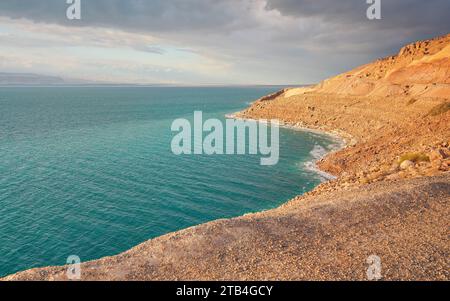 Rive de la mer morte du côté du Jourdain, plage de sable sec et rochers, le soleil brille sur la belle surface de l'eau azur Banque D'Images