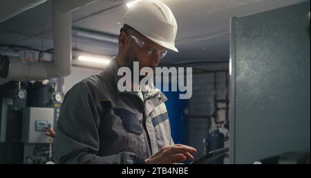 Ingénieur caucasien professionnel dans l'uniforme de sécurité et le casque de protection utilise l'ordinateur tablette numérique pour vérifier et ajuster le système d'énergie. Travailleur industriel travaille sur l'usine de fabrication moderne. Banque D'Images