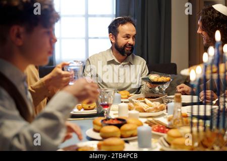 Concentrez-vous sur un homme juif barbu mature offrant à son fils ou à son invité d'essayer des pâtisseries maison traditionnelles tout en tenant une assiette avec de savoureux beignets Banque D'Images