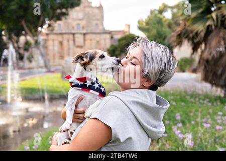 Femme avec les cheveux gris courts tenant et embrassé sur les lèvres par un petit chien senior dans le parc en plein air. Le chien porte le bandana avec un drapeau américain. Joie et compa Banque D'Images