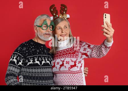 Couple senior en pulls de Noël, bandeau de renne et lunettes drôles prenant selfie sur fond rouge Banque D'Images