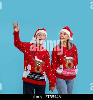 Couple senior dans des pulls de Noël et des chapeaux de Père Noël regardant quelque chose sur fond bleu clair. Espace pour le texte Banque D'Images