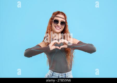 Élégante jeune femme hippie dans des lunettes de soleil faisant coeur avec les mains sur fond bleu clair Banque D'Images