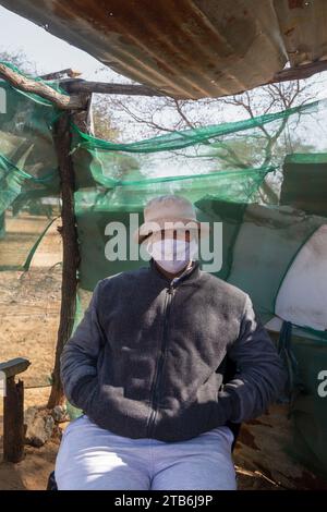 villageois jeune homme africain avec un chapeau portant un masque dans un bidonville situé sur une chaise à l'ombre d'une cabane Banque D'Images