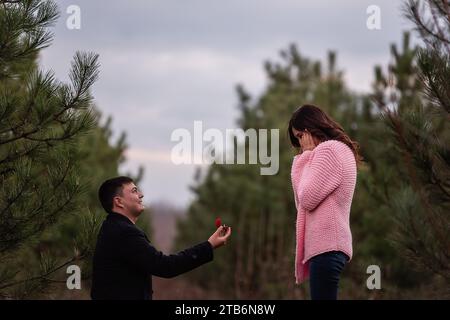 Jeune homme en manteau noir s'agenouille devant une belle femme, propose le mariage, tenant une boîte rouge avec bague en or dans la forêt. Fille en swea tricoté rose Banque D'Images