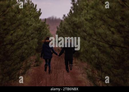 Heureux couple courant à travers la forêt se tenant la main. Un homme en manteau noir et une jeune femme en pull tricoté se baladent dans un parc de pins, pla Banque D'Images