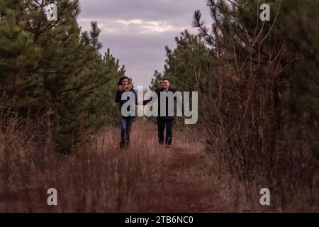 Heureux couple courant à travers la forêt se tenant la main. Un homme en manteau noir et une jeune femme en pull tricoté se baladent dans un parc de pins, pla Banque D'Images